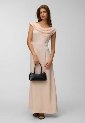 Jeune femme debout portant une robe sans manches rose pâle avec des détails en dentelle, tenant un petit sac à main noir et portant des sandales noires.