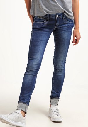 Osoba nosząca ciemnoniebieskie obcisłe jeansy z podwiniętymi nogawkami, szarą koszulkę i białe trampki, stojąca ze skrzyżowanymi nogami.