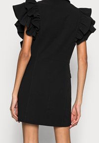 Svart minibalklänning med korta volangärmar, visad bakifrån där armarna hänger avslappnat vid sidorna.