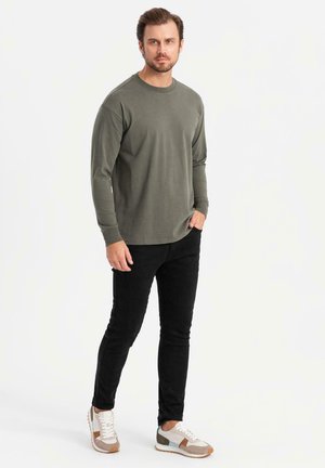 Hombre con camisa de manga larga color oliva, pantalones negros ajustados y zapatillas blancas, de pie con una mano en el bolsillo sobre un fondo blanco.