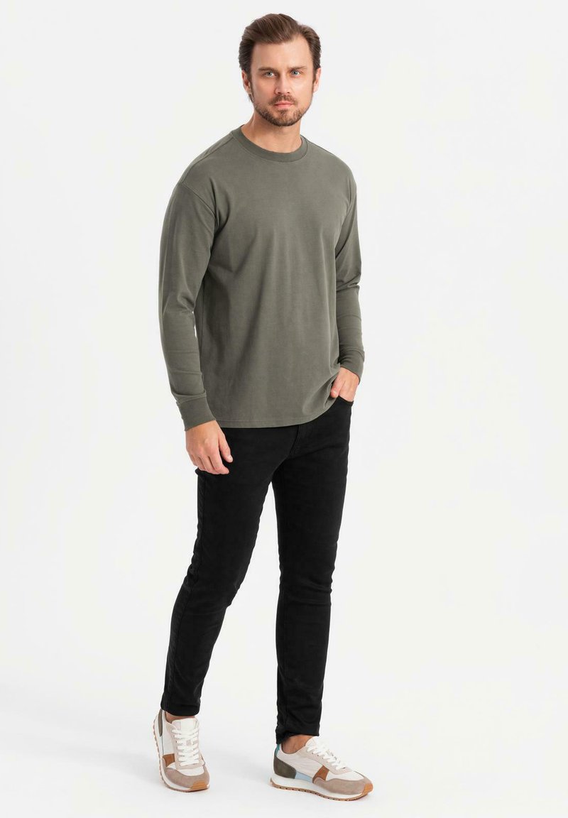 Mies, joka on pukeutunut oliivinvihreään pitkähihaiseen paitaan, mustiin slim-fit-housuihin ja valkoisiin lenkkareihin, seisoo yksi käsi taskussa valkoista taustaa vasten.