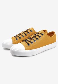 Žluté plátěné tenisky s bílým gumovým špičkou a podrážkou, černými tkaničkami a oky, a texturovaným svrchním povrchem.