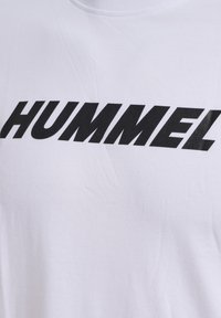 Hvid bomulds t-shirt med et stort sort logo tekst "HUMMEL" i fed, vinklet skrifttype på brystområdet. Glat tekstur.