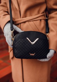 Schwarze, gesteppte Handtasche mit einem goldenen Akzent und Logo, ausgestattet mit einem Reißverschluss und einem abnehmbaren Schultergurt, gehalten von einer behandschuhten Hand.