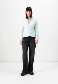 Vrouw die tegen een witte achtergrond staat, gekleed in een lichtblauwe gewatteerde jas, zwarte wijde jeans en zwarte plateau schoenen.