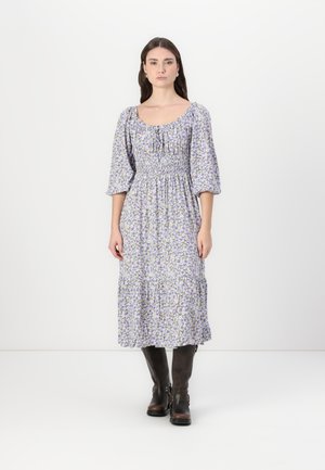 Blumen-Midi-Kleid mit Puffärmeln, lila und gelben Mustern auf weißem Hintergrund, geraffter Taille und gestuftem Rock. Getragen mit dunklen Stiefeln.