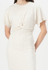 Knot-front mekko vaaleassa teksturoitussa kankaassa, jossa on lyhyet hihat, mukautuva siluetti ja hienovarainen kudontakuvi. 