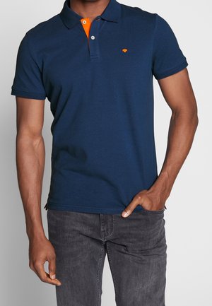 Mand iført en navyblå poloshirt med orange indvendig krave og lille orange logo, kombineret med mørkegrå jeans, hånd i lommen.