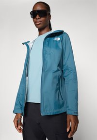 Teal värvitud tuulejope, millel on tõmblukuga eesmine osa, reguleeritav kapuuts ja taskud. Koos helesinise ülemise ja mustade pükstega, mille pind on sile.