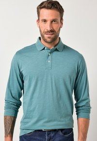 Polo shirt turchese a maniche lunghe con colletto e tre bottoni, indossato su jeans in denim scuro. Tessuto morbido, design informale.