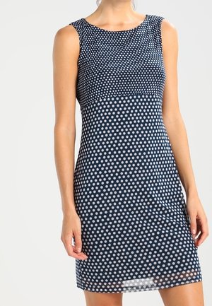 Granatowa sukienka bez rękawów w białe kropki, dopasowana w talii, o długości do kolan, noszona przez stojącą osobę na jednolitym tle.