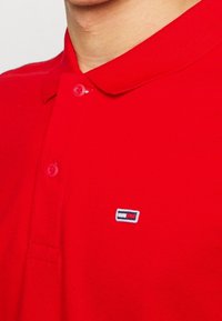 Červené polo tričko vyrobené z bavlny, s klasickým límečkem, dvěma knoflíky a malým vyšitým logem v modré, bílé a červené barvě na hrudi.
