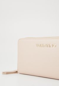 Rožinis Valentino piniginė su tekstūruotu paviršiumi, su auksiniu logotipu ir užtrauktuku. Kompaktiška forma su šonine anga patogiam laikymui.