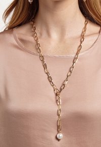 Collar de eslabones de oro con un gran colgante de perla lisa, con eslabones de cadena alargados y un diseño en forma de Y, llevado sobre una blusa rosa clara.