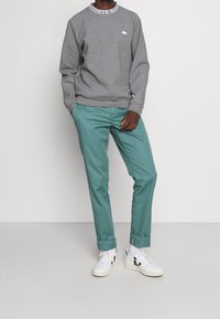 Person som bär en grå Lacoste-tröja med logotyp på kragen och teal-uppvikt byxor, stående mot en enkel ljus bakgrund.