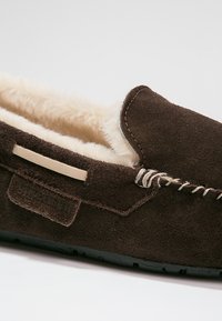 Pantofole mocassini in suede marrone con fodera in shearling color crema, cuciture decorative e un accento beige sul lato. Suola in gomma nera.