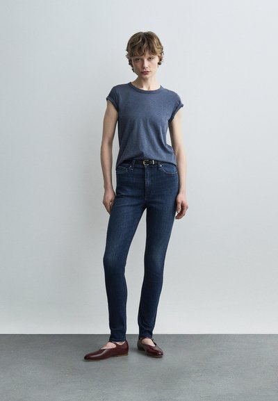 Νεαρή γυναίκα που στέκεται μπροστά σε έναν ανοιχτό χρωματικά τοίχο, φοράει μπλε μπλούζα με γυρισμένα μανίκια, σκουρόχρωμο στενό τζιν, μαύρη ζώνη και μπορντό επίπεδα παπούτσια.