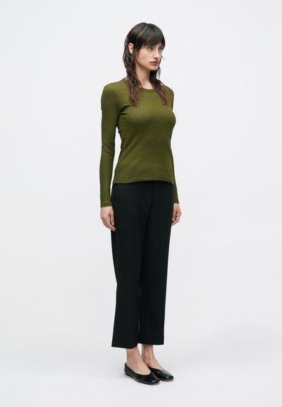 Μπλούζα με μακριά μανίκια και ριμπ ύφανση σε σκούρο πράσινο, συνδυασμένη με μαύρο παντελόνι φαρδύ στην ποδιά και μαύρα slip-on παπούτσια, στιλιζαρισμένη σε μινιμαλιστικό στυλ.