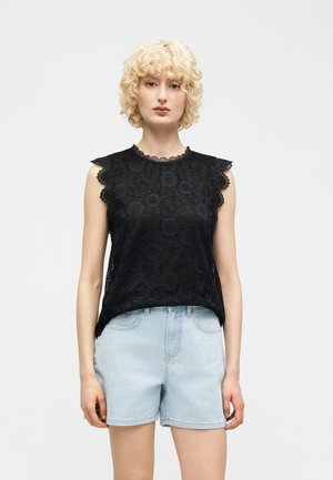 Kobieta o kręconych blond włosach, ubrana w czarny koronkowy top bez rękawów i jasnoniebieskie szorty jeansowe, stojąca na jednolitym białym tle.