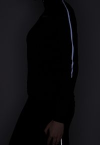 Svart långärmad topp med hög krage, prydd med en reflekterande vit rand längs axeln, mot en mörk bakgrund.