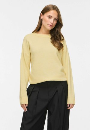 Femme aux longs cheveux bruns porte un pull léger jaune à manches longues et un pantalon noir plissé taille haute sur fond blanc.