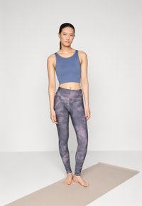 Blauer gerippter Cropped Top kombiniert mit lila grafischen Leggings, die ein strukturiertes Muster aufweisen, beide aus dehnbarem Stoff, auf einer Yogamatte.