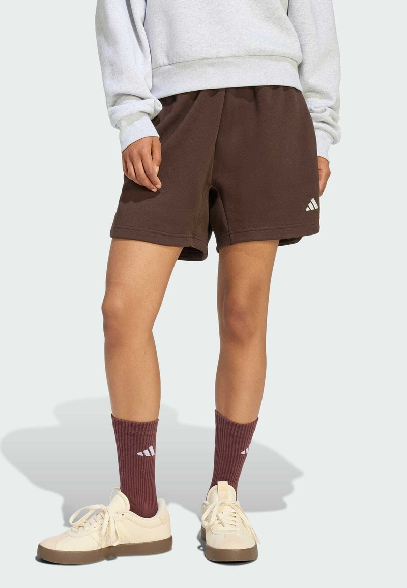 Osoba v hnědých šortkách Adidas, odpovídajících hnědých ponožkách Adidas, krémových teniskách a světle šedé mikině stojící na jednoduchém pozadí.