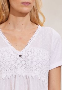 Femme portant une blouse blanche avec un motif floral en dentelle et une texture à pois, mettant en valeur le décolleté et la partie supérieure de la poitrine.