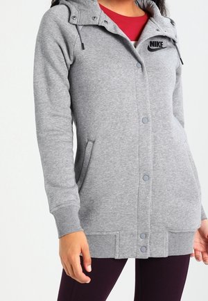 Osoba nosząca szarą bluzę z kapturem Nike z przednimi zatrzaskami, bocznymi kieszeniami i kapturem, na czerwonym topie i ciemnych spodniach.
