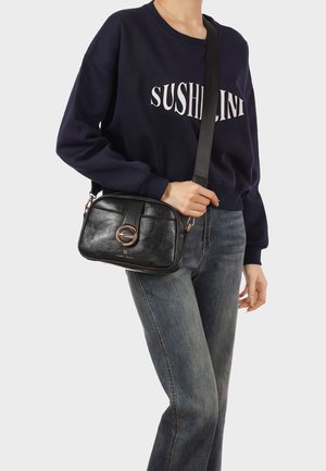 Persona che indossa una felpa blu navy con la scritta "SUSHILANI" e jeans blu, che tiene una borsa a tracolla nera in pelle con chiusura circolare dorata.