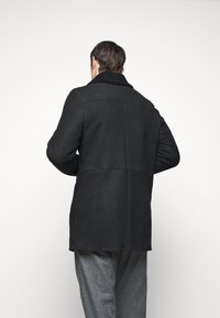 Manteau en daim noir avec un col en laine, coupe droite et des détails de couture, associé à un pantalon gris. La vue arrière montre une texture lisse.