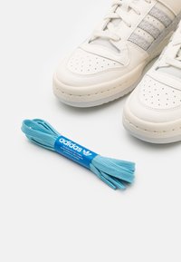 Białe skórzane buty z szarymi akcentami i perforowanymi detalami, w opakowaniu z niebieskimi sznurówkami i logo Adidas.