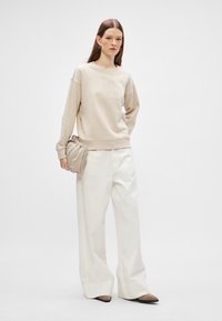 Beżowy sweter z wypukłym wzorem, w połączeniu z szerokimi, białymi spodniami. Modelka trzyma fakturowaną beżową torebkę. Buty są dwukolorowe z ostro zakończonymi noskami.