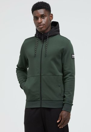 Kapuzenjacke mit Reißverschluss in Dunkelgrün mit einer schwarzen Innenseite der Kapuze, ausgestattet mit Kordelzügen, zwei Fronttaschen und einem seitlichen Logopatch.