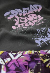 Un tissu noir arbore un texte graphique vibrant en violet et rose "Déploie tes ailes" au-dessus d'un motif floral en jaune et violet.