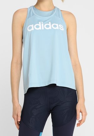 Kvinde iført lyseblå Adidas ærmeløs top og sorte leggings med mønster, stående med armene afslappet ned langs siderne mod en ensfarvet baggrund.
