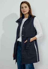 Marinblå ärmlös jacka med dragkedja och tryckknappar, sidofickor, bälte och reflexdetaljer. Bärs över en ljus topp och jeans.