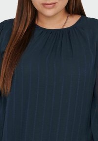 Mörkblå blus med vertikala ränder, mjukt tyg, rynkad halsringning och långa ärmar med genomskinliga detaljer. Tätt sittande design.
