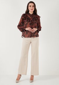 Sheer, patterned blouse in rust and black with ruffled collar and cuffs, paired with cream wide-leg trousers and metallic heels.