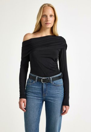 Kvinde iført sort off-shoulder bluse med lange ærmer og blå jeans med sort bælte, der står mod en ensfarvet lys baggrund.