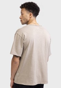 Tricou bej cu mânecă scurtă, cu o croială lejeră, margini brut din material texturat. O etichetă mică cu logo este poziționată pe cusătura laterală.