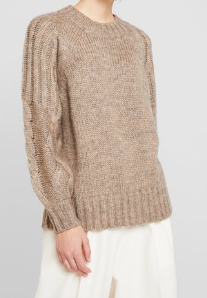 Kraftig beige kabelstrikket sweater med ribbet manchetter og kant, båret med løse hvide bukser på en person mod en ensfarvet baggrund.