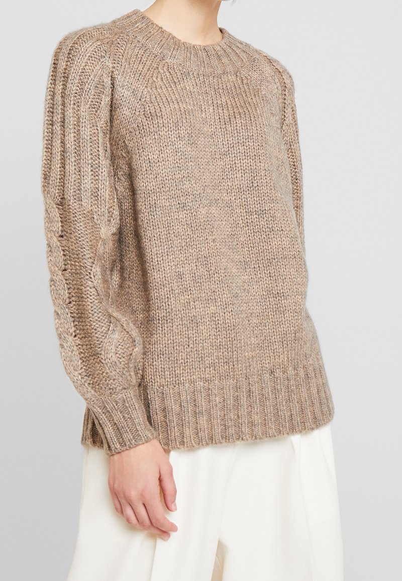 Pull épais beige en maille torsadée avec poignets et ourlet côtelés, porté avec un pantalon blanc ample sur une personne devant un fond uni.