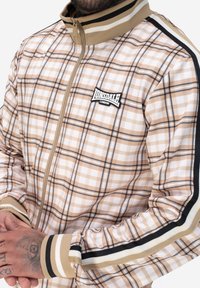 Kariertes Zip-Jackett in Beigetönen mit Schwarz- und Weißakzenten. Verfügt über einen Stehkragen und elastische Bündchen mit gestreiften Details.