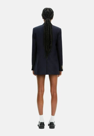 Blazer navy con spalle strutturate, singolo spacco posteriore e finiture con bottoni dorati. Indossato con calzini bianchi e scarpe nere.