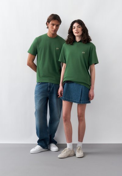 Мъж и жена стоят един до друг, облечени в еднакви зелени тениски на Lacoste с лого, жената е с синя дънкова пола и маратонки, мъжът е с сини дънки и бели обувки.