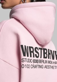 Felpa con cappuccio rosa dalla texture morbida. Presenta un testo stampato in nero sulla schiena, che mostra dettagli del marchio e del design.