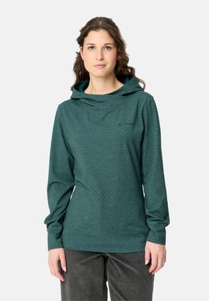 Frau mit welligem braunem Haar, die ein grünes langärmeliges Kapuzenshirt und dunkle Hose trägt und vor einem schlichten hellen Hintergrund steht.