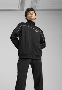 Chándal deportivo negro hecho de tejido suave, con detalles en blanco y una chaqueta con cremallera y cuello alto. El logotipo se muestra de manera prominente.