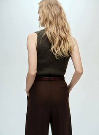 Ärmelloses, dunkelgrünes Stricktop, kombiniert mit hochgeschnittenen braunen Hosen und einem braunen Gürtel. Das Haar ist wellig und fällt über die Schultern.